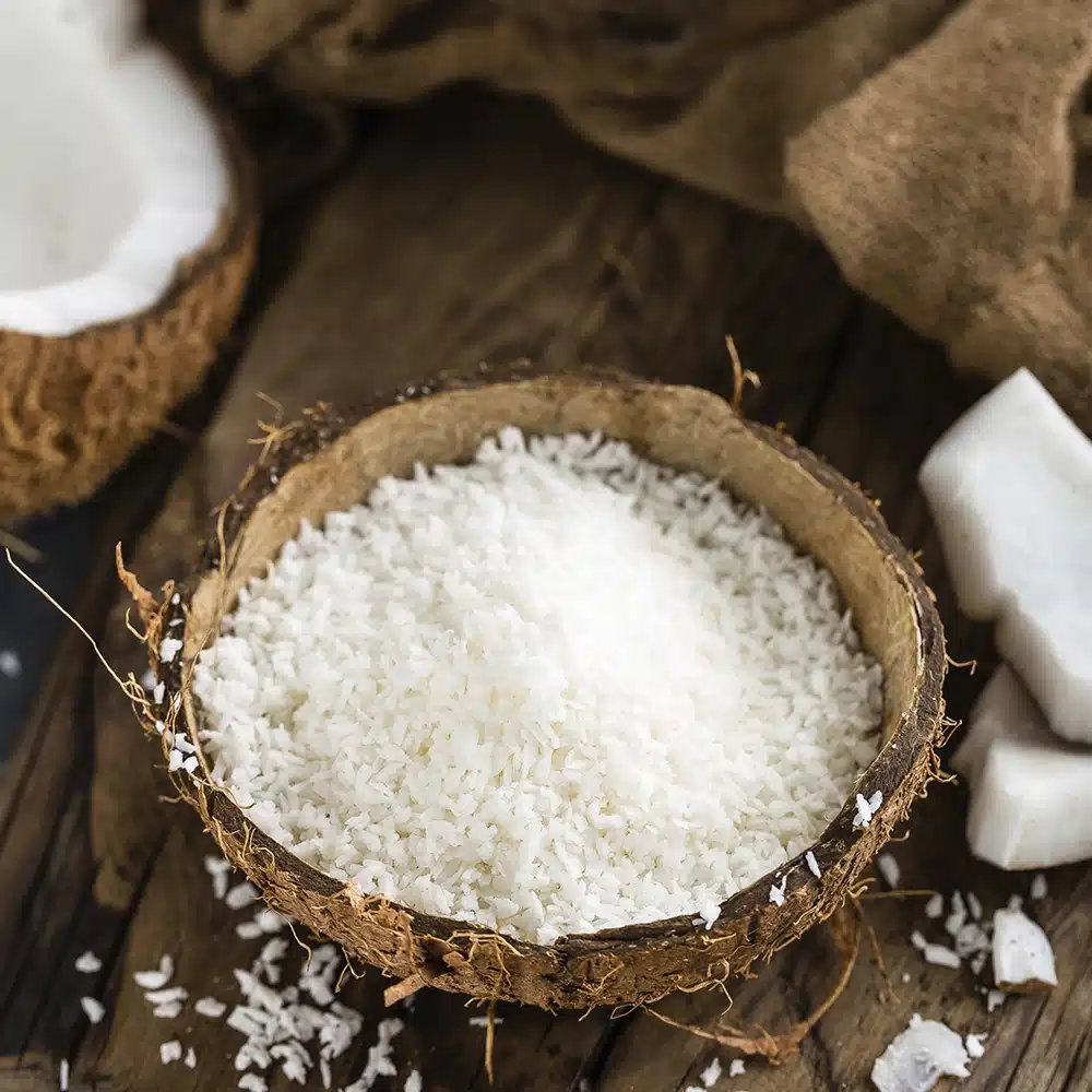 A bowl of desiccated coconut on a wooden surface with coconut pieces around.