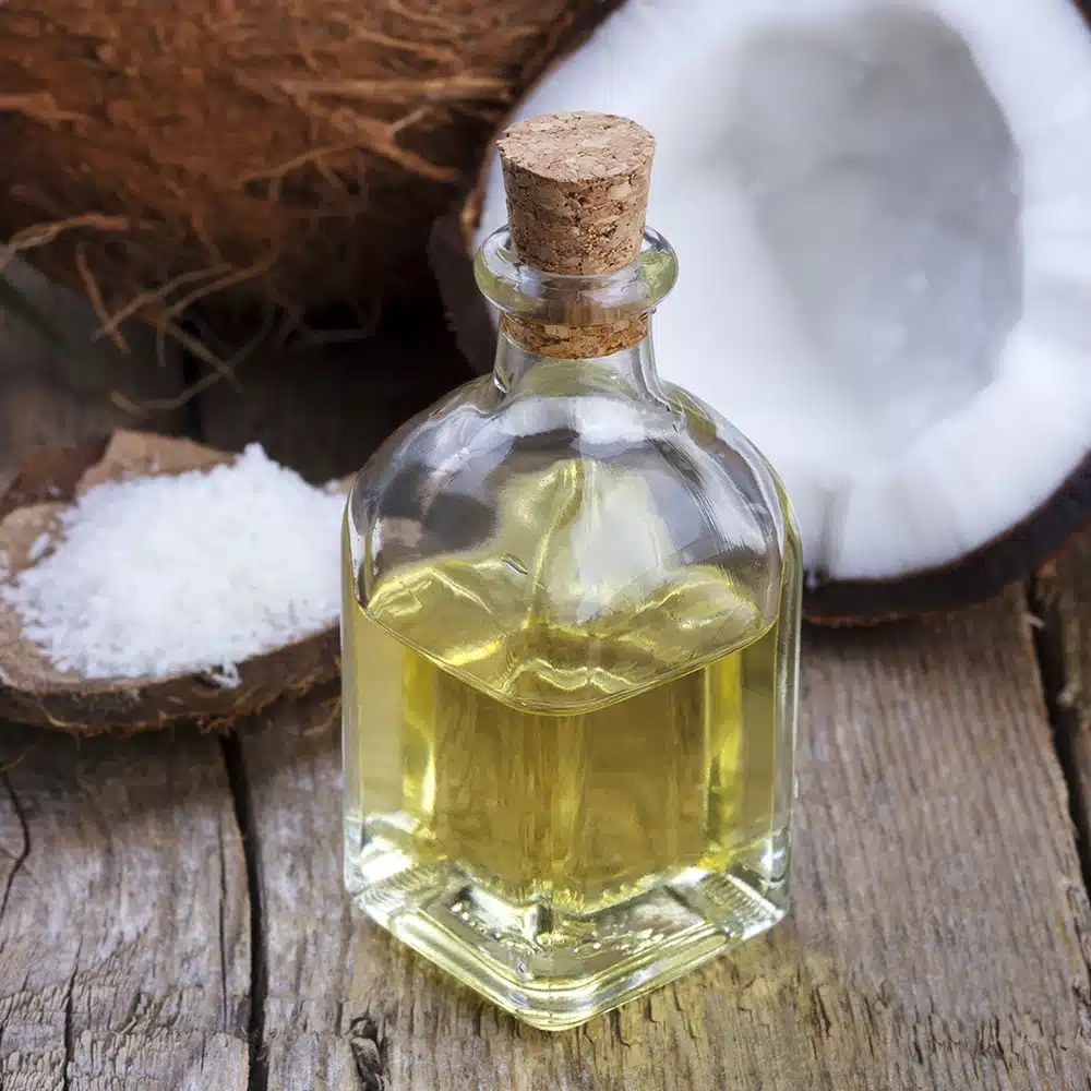 Bottle of refined coconut oil with coconut halves and shredded coconut on a wooden surface.