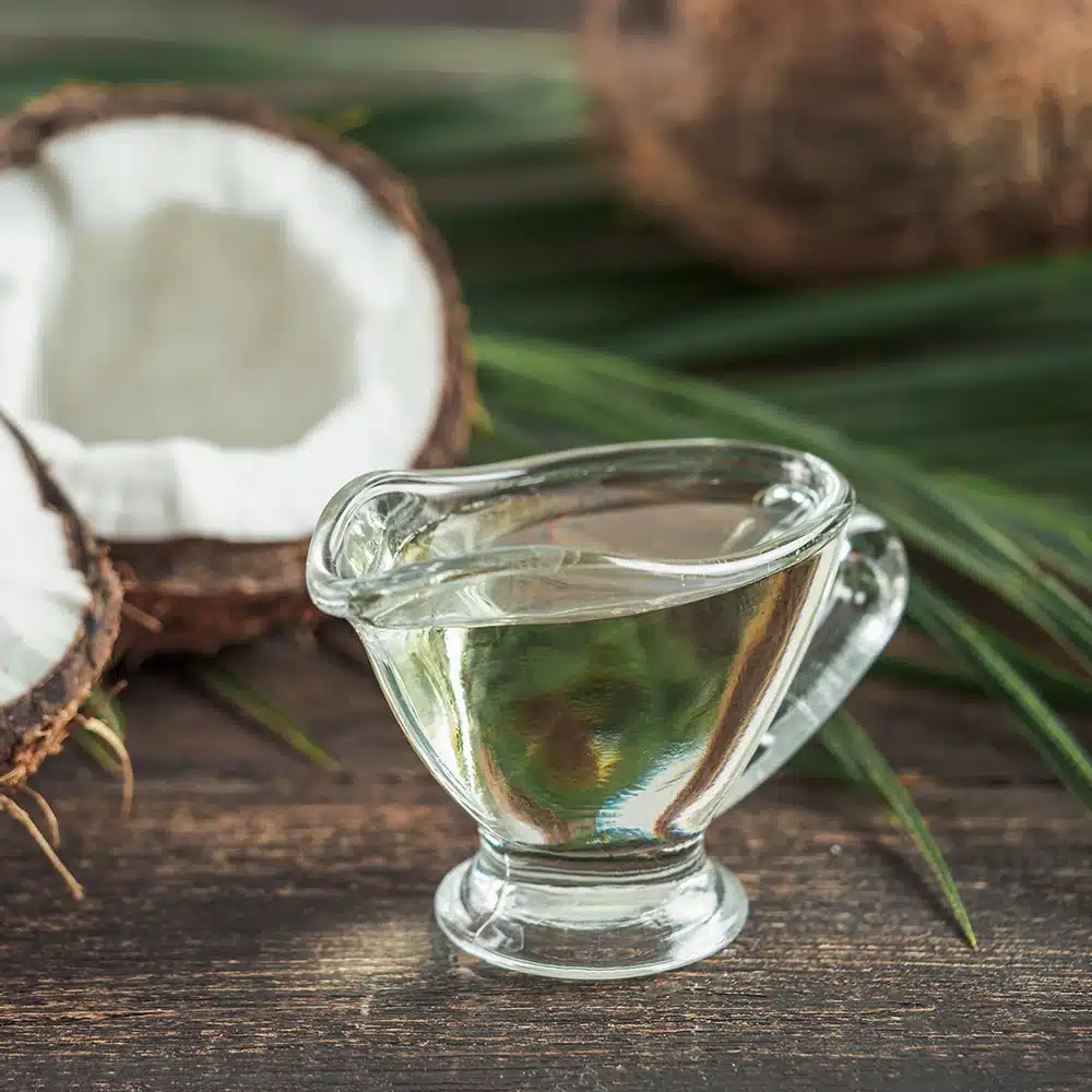 Glass pitcher filled with virgin coconut oil with coconut halves and palm leaves in the background.